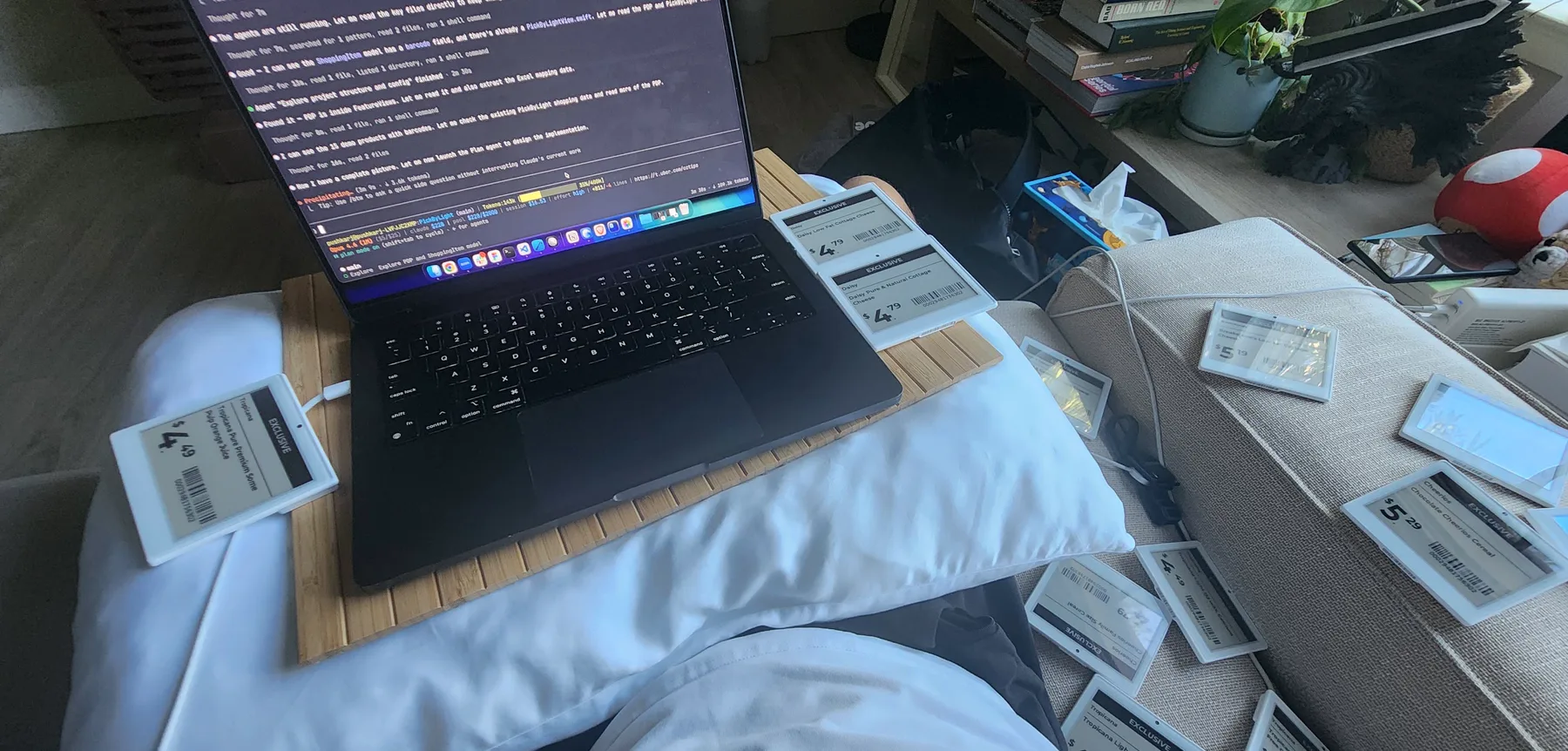 A laptop on a lap desk, surrounded by a dozen electronic shelf labels showing prices and barcodes